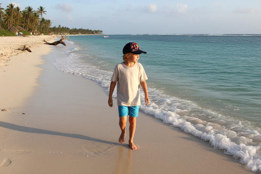 May include: A person wearing a light-colored t-shirt, blue shorts, and a baseball cap walks along a sandy beach. The ocean is visible in the background, with palm trees and a clear sky.