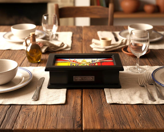May include: A black wooden box with a colorful Guatemalan design on top sits on a wooden table set for a meal. The table is set with plates, silverware, glasses, and a small bottle of oil. The box has the word "GUATEMALA" on it.